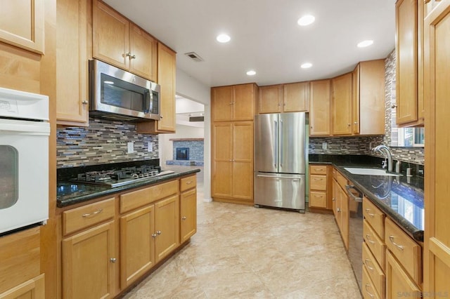 kitchen with a fireplace, dark stone countertops, decorative backsplash, sink, and stainless steel appliances