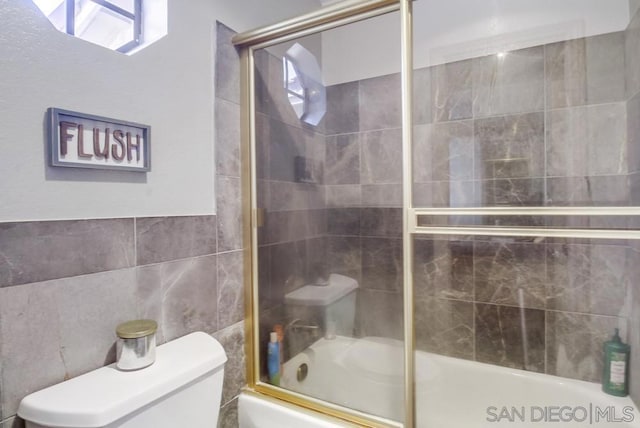 bathroom featuring tile walls, combined bath / shower with glass door, and toilet