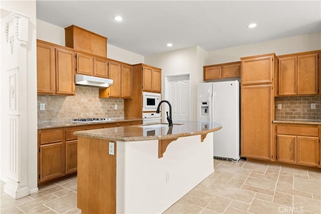 kitchen featuring sink, light stone counters, white appliances, a kitchen island with sink, and backsplash