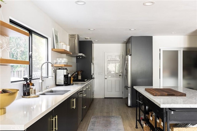 kitchen with dark wood-type flooring, wall chimney range hood, stovetop, sink, and stainless steel refrigerator