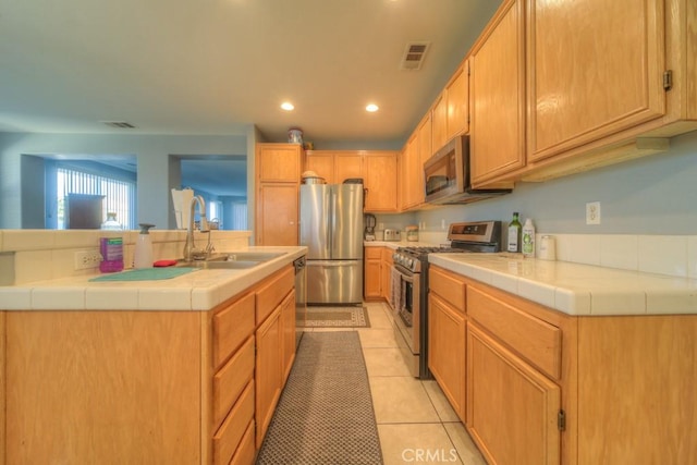 kitchen with light tile patterned floors, stainless steel appliances, tile counters, a kitchen island with sink, and sink