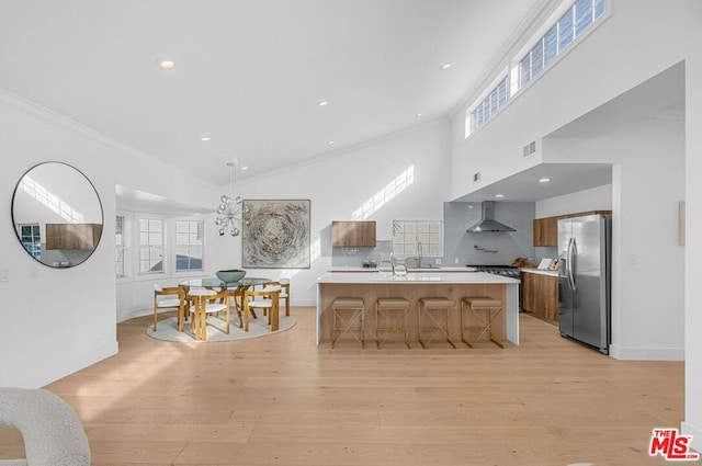 kitchen with stainless steel refrigerator with ice dispenser, wall chimney exhaust hood, a kitchen bar, a center island with sink, and ornamental molding