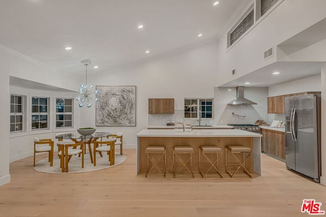 kitchen with pendant lighting, a kitchen bar, stainless steel refrigerator with ice dispenser, a center island with sink, and wall chimney exhaust hood