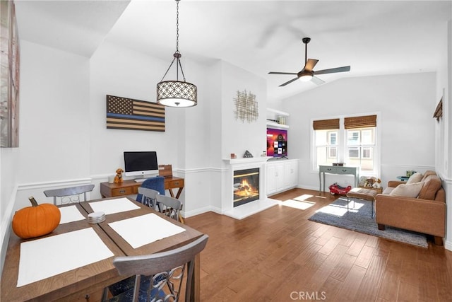 living area featuring lofted ceiling, a glass covered fireplace, wood finished floors, baseboards, and ceiling fan