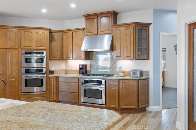 kitchen featuring exhaust hood, stainless steel appliances, decorative backsplash, light hardwood / wood-style flooring, and light stone counters