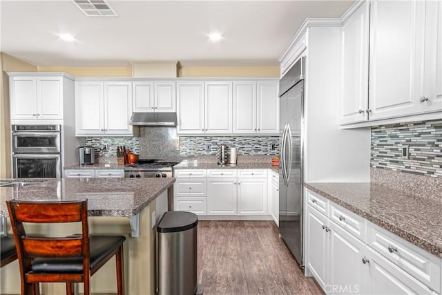 kitchen featuring white cabinets, appliances with stainless steel finishes, wood-type flooring, stone counters, and a breakfast bar area