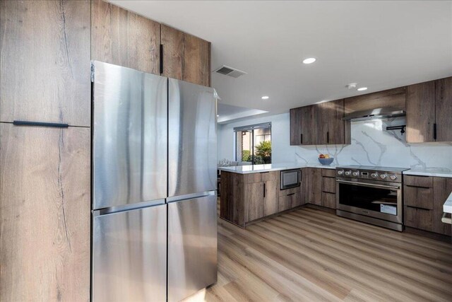 kitchen featuring light hardwood / wood-style floors, kitchen peninsula, appliances with stainless steel finishes, backsplash, and wall chimney range hood