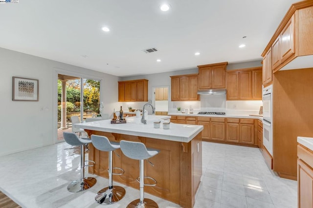 kitchen featuring white double oven, a center island with sink, stainless steel gas cooktop, a breakfast bar, and sink