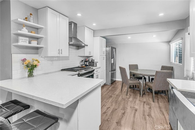 kitchen with white cabinetry, kitchen peninsula, appliances with stainless steel finishes, light wood-type flooring, and wall chimney exhaust hood