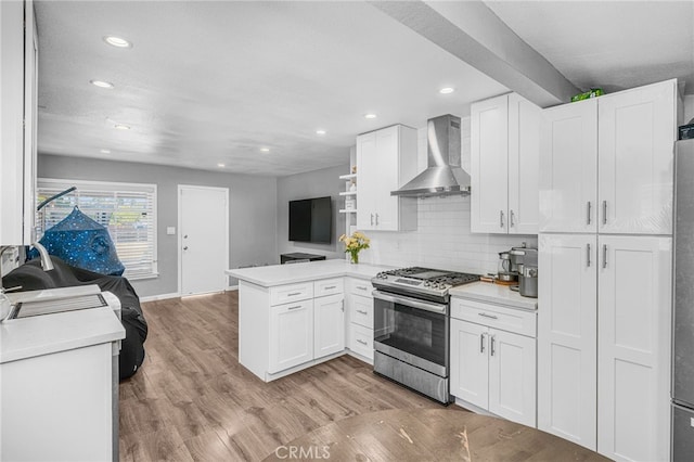 kitchen featuring light hardwood / wood-style floors, kitchen peninsula, stainless steel range with gas cooktop, white cabinets, and wall chimney exhaust hood