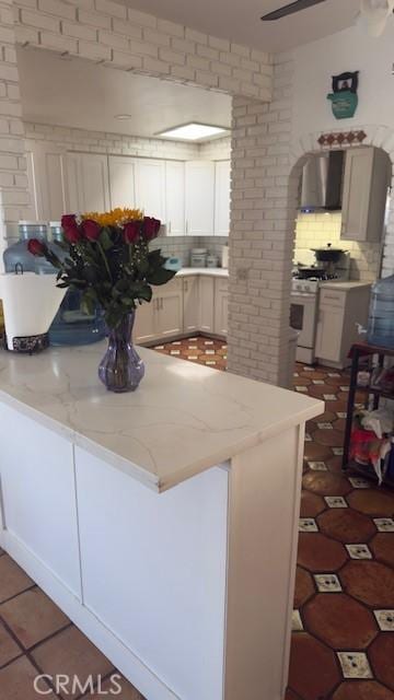 kitchen with white gas range oven, kitchen peninsula, white cabinetry, a kitchen breakfast bar, and wall chimney exhaust hood