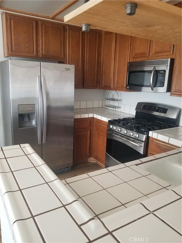 kitchen with tile counters, appliances with stainless steel finishes, and brown cabinetry