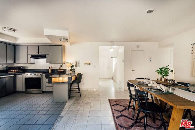 kitchen with a kitchen bar, tasteful backsplash, gray cabinetry, stainless steel appliances, and range hood