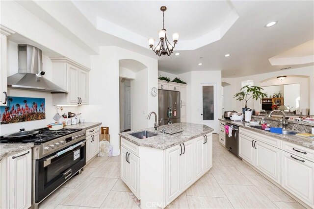 kitchen featuring a raised ceiling, sink, a kitchen island with sink, stainless steel appliances, and wall chimney exhaust hood