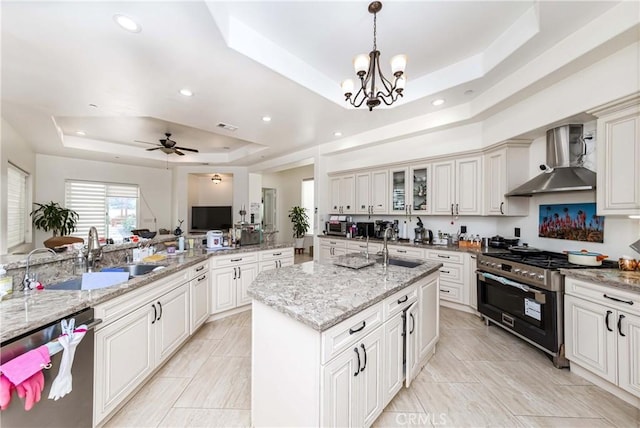 kitchen featuring an island with sink, stainless steel appliances, a raised ceiling, wall chimney range hood, and ceiling fan with notable chandelier