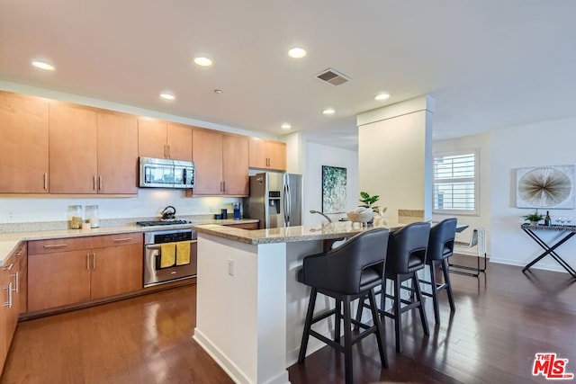 kitchen featuring a breakfast bar, a kitchen island with sink, dark wood-type flooring, stainless steel appliances, and light stone counters