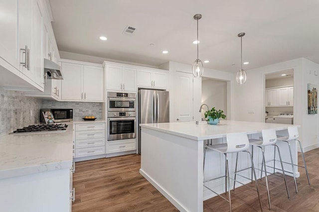 kitchen with appliances with stainless steel finishes, an island with sink, white cabinets, backsplash, and hanging light fixtures