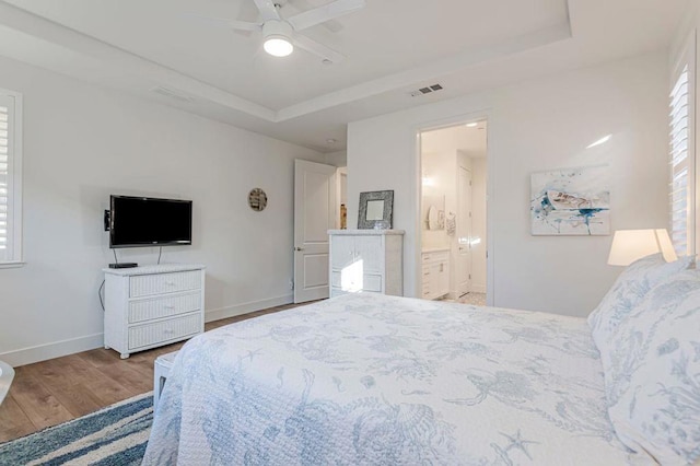 bedroom featuring ensuite bathroom, multiple windows, ceiling fan, light hardwood / wood-style floors, and a raised ceiling