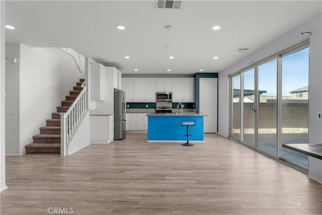 kitchen with white cabinetry, light wood-type flooring, dark stone countertops, stainless steel appliances, and a kitchen island with sink