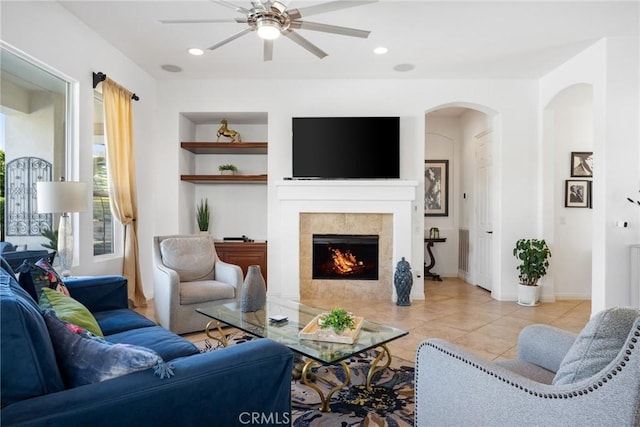 living room featuring ceiling fan, light tile patterned floors, built in features, and a tiled fireplace