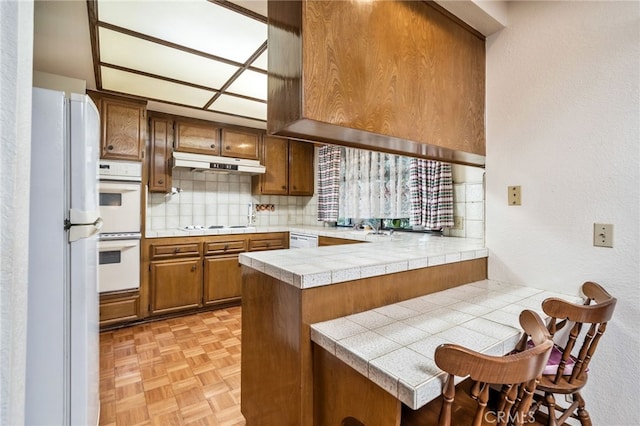 kitchen featuring light parquet floors, kitchen peninsula, tile counters, decorative backsplash, and white appliances