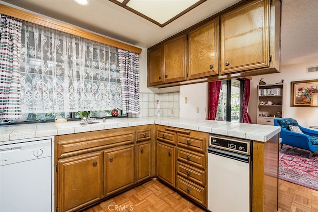 kitchen featuring tile countertops, kitchen peninsula, backsplash, white dishwasher, and light parquet flooring