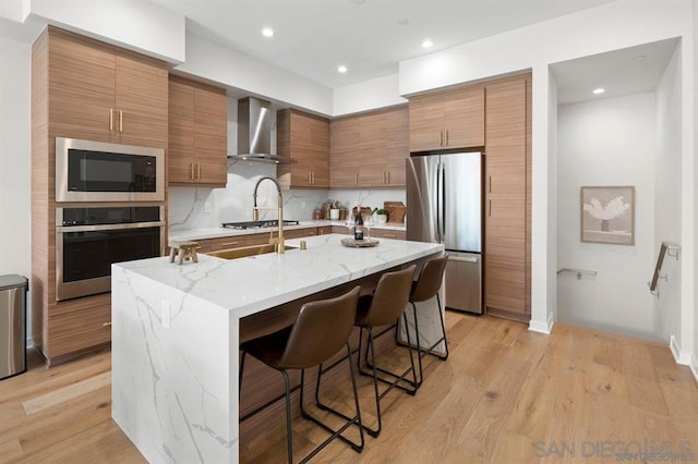 kitchen with a kitchen bar, a kitchen island with sink, appliances with stainless steel finishes, wall chimney exhaust hood, and light stone counters