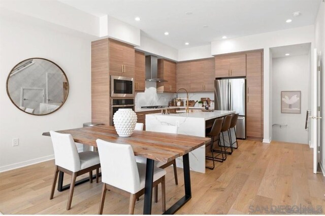 kitchen featuring tasteful backsplash, wall chimney range hood, light wood-type flooring, appliances with stainless steel finishes, and a kitchen breakfast bar