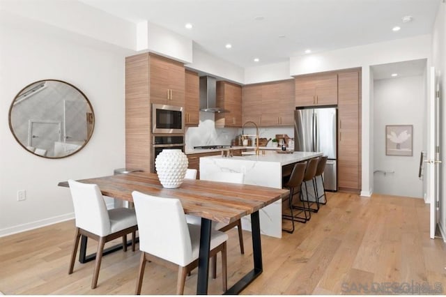 kitchen with appliances with stainless steel finishes, wall chimney exhaust hood, backsplash, a breakfast bar, and light hardwood / wood-style flooring