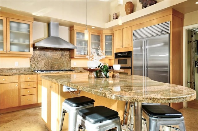 kitchen featuring decorative light fixtures, a kitchen breakfast bar, stainless steel appliances, light stone countertops, and wall chimney range hood