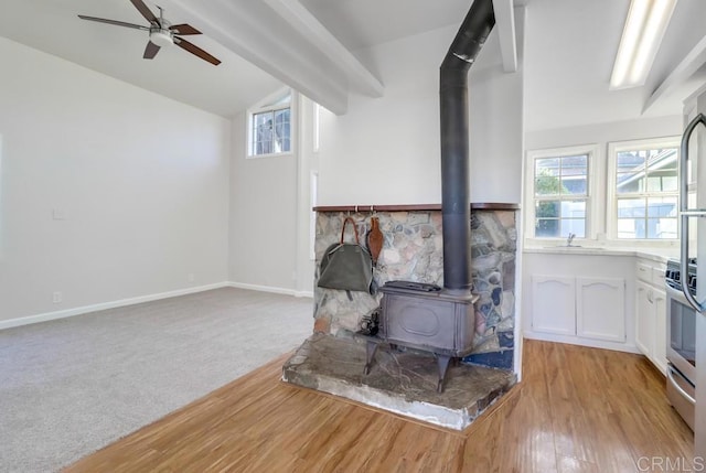 carpeted living room with ceiling fan, a wood stove, sink, and vaulted ceiling with beams