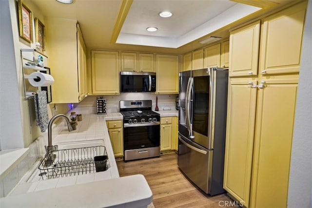 kitchen featuring tile countertops, a raised ceiling, sink, light hardwood / wood-style flooring, and appliances with stainless steel finishes