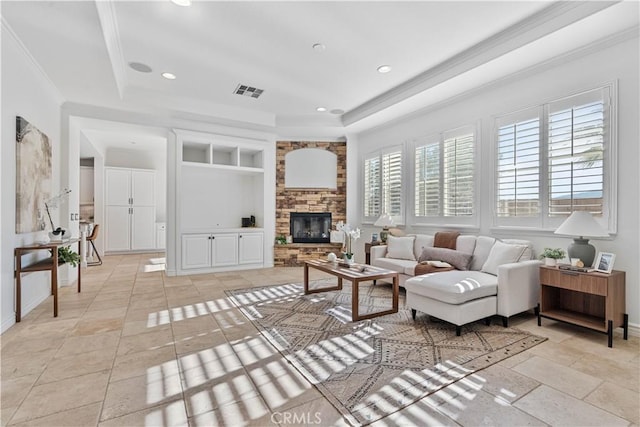 living room with a raised ceiling, built in shelves, crown molding, and a stone fireplace