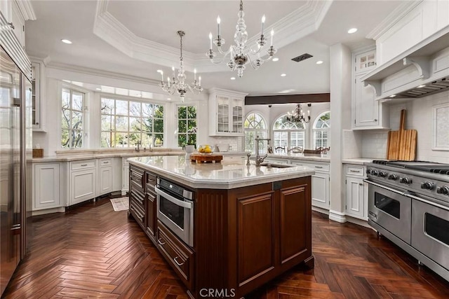 kitchen with dark parquet floors, a center island with sink, a raised ceiling, and stainless steel appliances