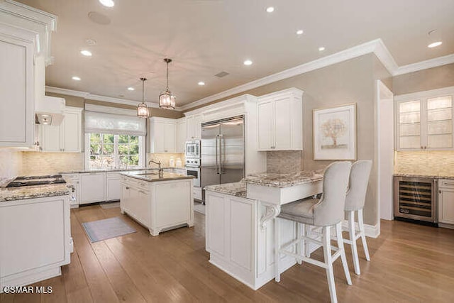 kitchen featuring wine cooler, a center island with sink, built in appliances, hanging light fixtures, and white cabinets