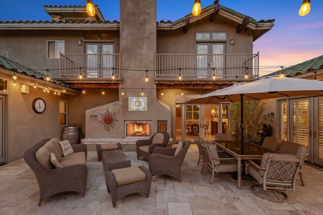 back house at dusk with a balcony, a patio area, and an outdoor living space with a fireplace