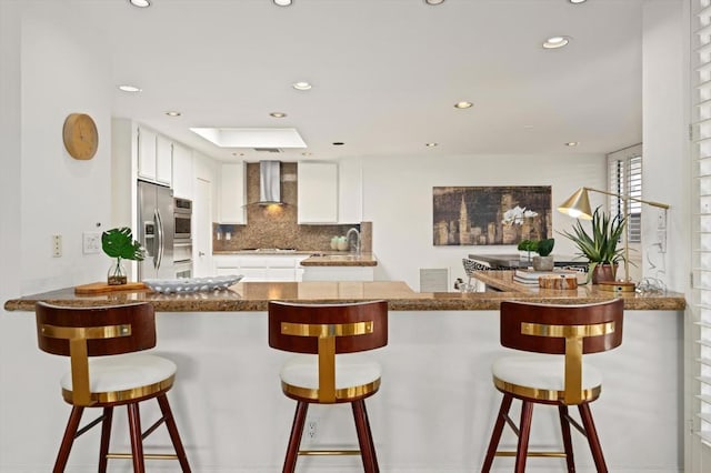 kitchen featuring stainless steel refrigerator with ice dispenser, wall chimney range hood, white cabinetry, a kitchen breakfast bar, and backsplash