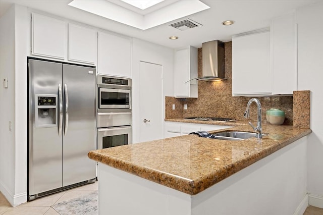 kitchen featuring wall chimney range hood, kitchen peninsula, light tile patterned flooring, stainless steel appliances, and white cabinets