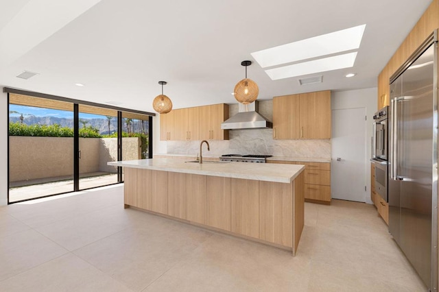 kitchen featuring light brown cabinetry, a skylight, hanging light fixtures, a kitchen island with sink, and wall chimney range hood