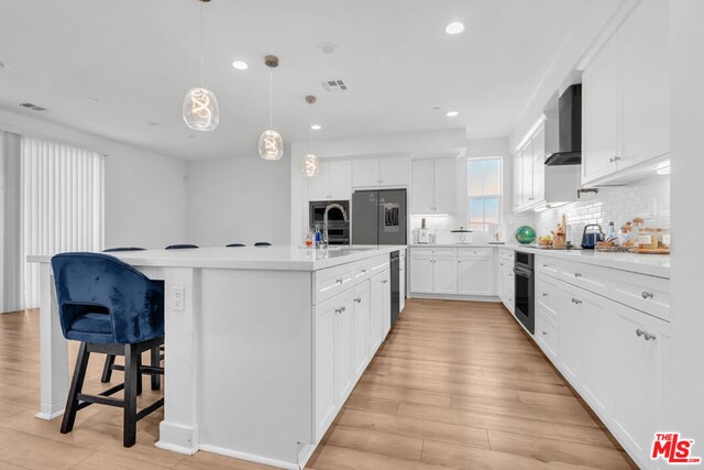 kitchen with wall chimney range hood, hanging light fixtures, a kitchen island with sink, stainless steel appliances, and white cabinets