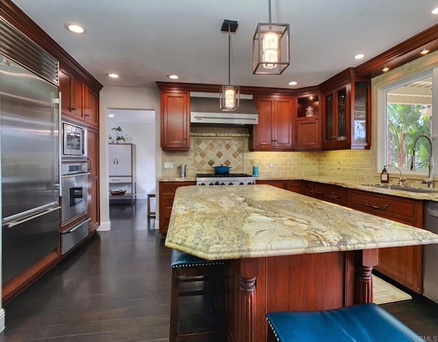 kitchen with decorative light fixtures, wall chimney range hood, a kitchen island, sink, and built in appliances