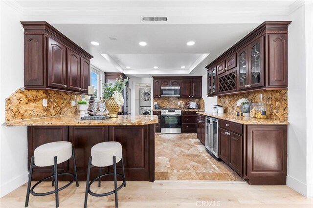 interior space featuring backsplash, a raised ceiling, kitchen peninsula, stacked washing maching and dryer, and appliances with stainless steel finishes