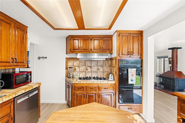 kitchen with under cabinet range hood, backsplash, appliances with stainless steel finishes, light countertops, and a wood stove