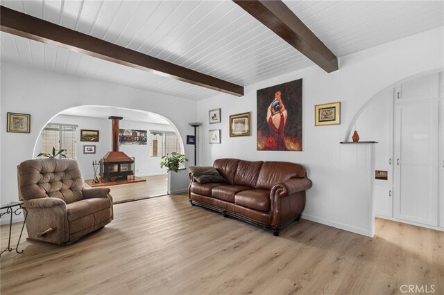 living area featuring arched walkways, beam ceiling, light wood-style floors, and a wood stove