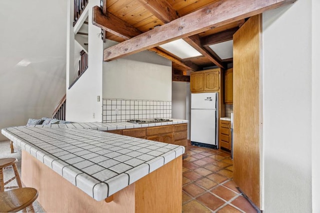 kitchen featuring stainless steel gas stovetop, tile countertops, backsplash, white refrigerator, and beam ceiling