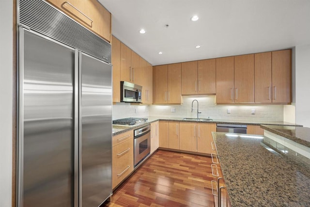 kitchen featuring appliances with stainless steel finishes, decorative backsplash, dark wood-type flooring, dark stone countertops, and sink