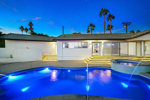 pool at dusk with pool water feature, a patio area, and an in ground hot tub