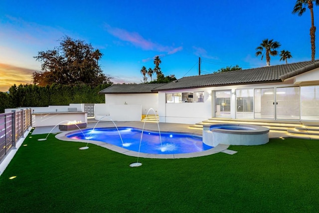 pool at dusk with pool water feature, an in ground hot tub, and a yard