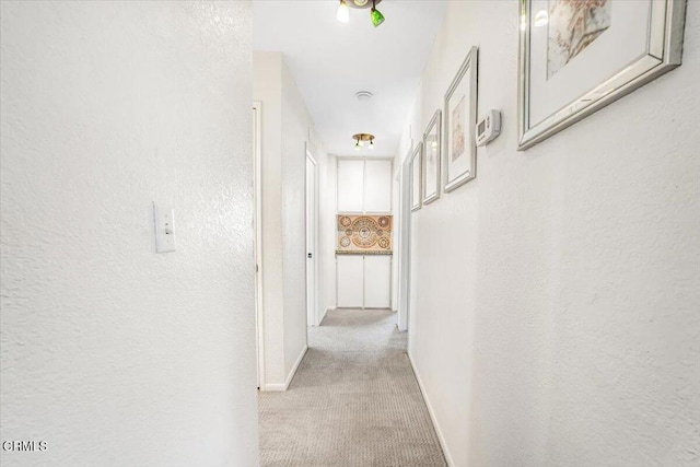 hall featuring light carpet, a textured wall, and baseboards