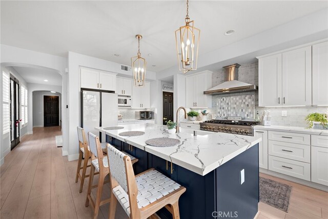 kitchen with light stone countertops, white appliances, wall chimney exhaust hood, white cabinetry, and an island with sink